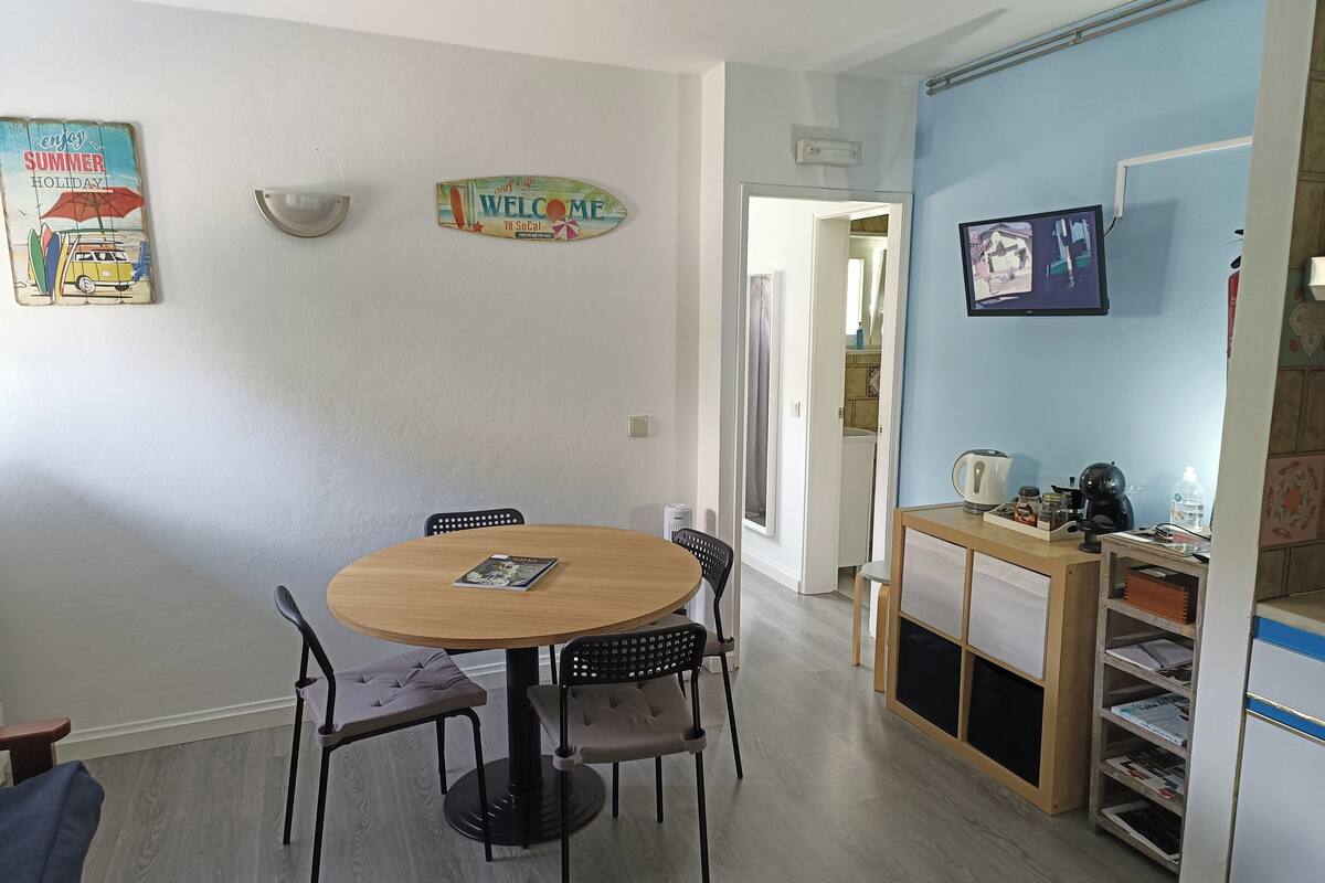 Un comedor con una mesa redonda y cuatro sillas, una pequeña cocina con cafetera, y un televisor montado en la pared.