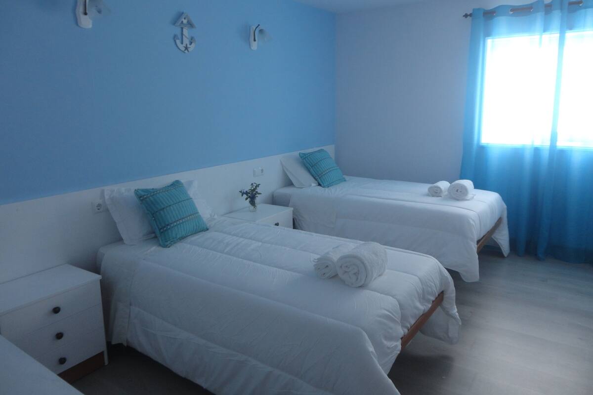 Habitación de hotel con dos camas, cada una con ropa blanca y almohada azul. Hay apliques de luz en la pared y una ventana con cortinas azules.