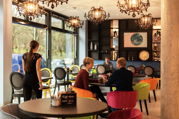 Un café moderno con mesas redondas y sillas coloridas, ventanas amplias y candelabros decorativos.