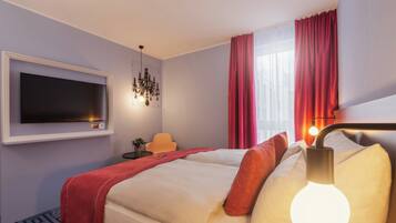 Une chambre d’hôtel avec un lit, un téléviseur fixé au mur, un lustre et une fenêtre avec des rideaux.