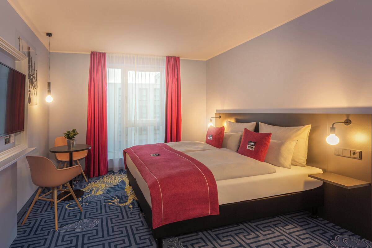 Ett hotellrum med en säng, ett skrivbord, en stol, en tv och ett fönster med gardiner.