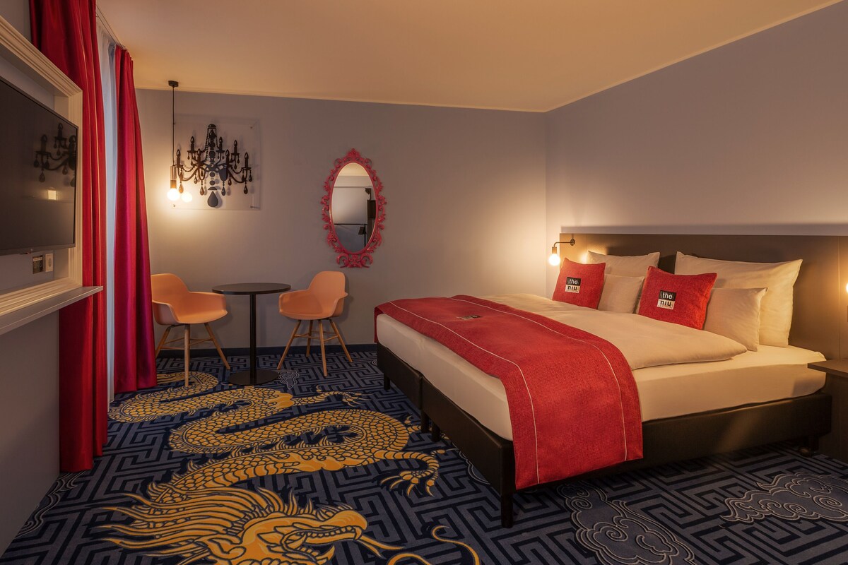 Habitación de hotel con cama, escritorio, dos sillas, televisor, espejo y candelabro.