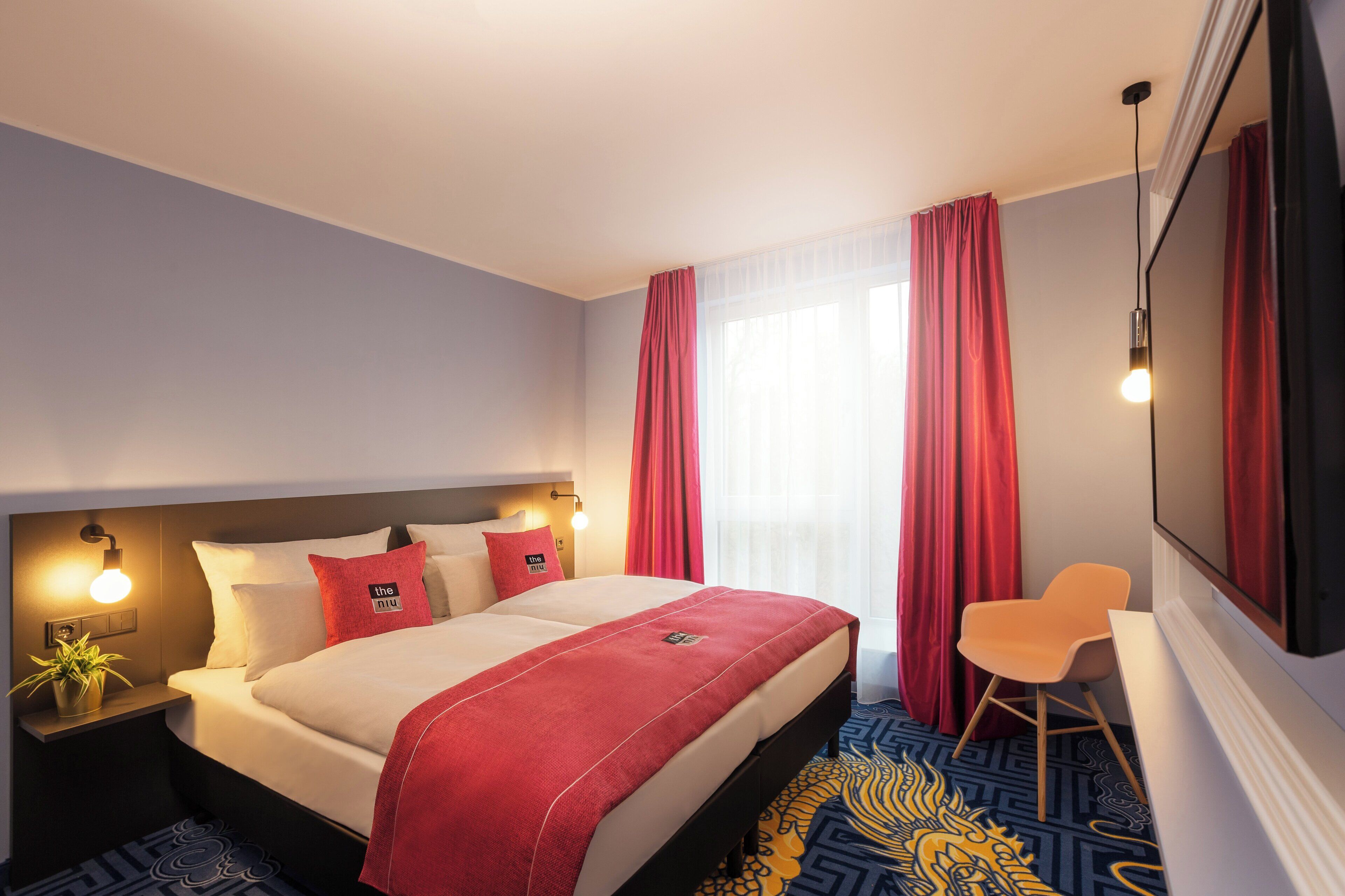 Habitación de hotel con cama, mesita de noche, silla, televisor y ventana con cortinas.