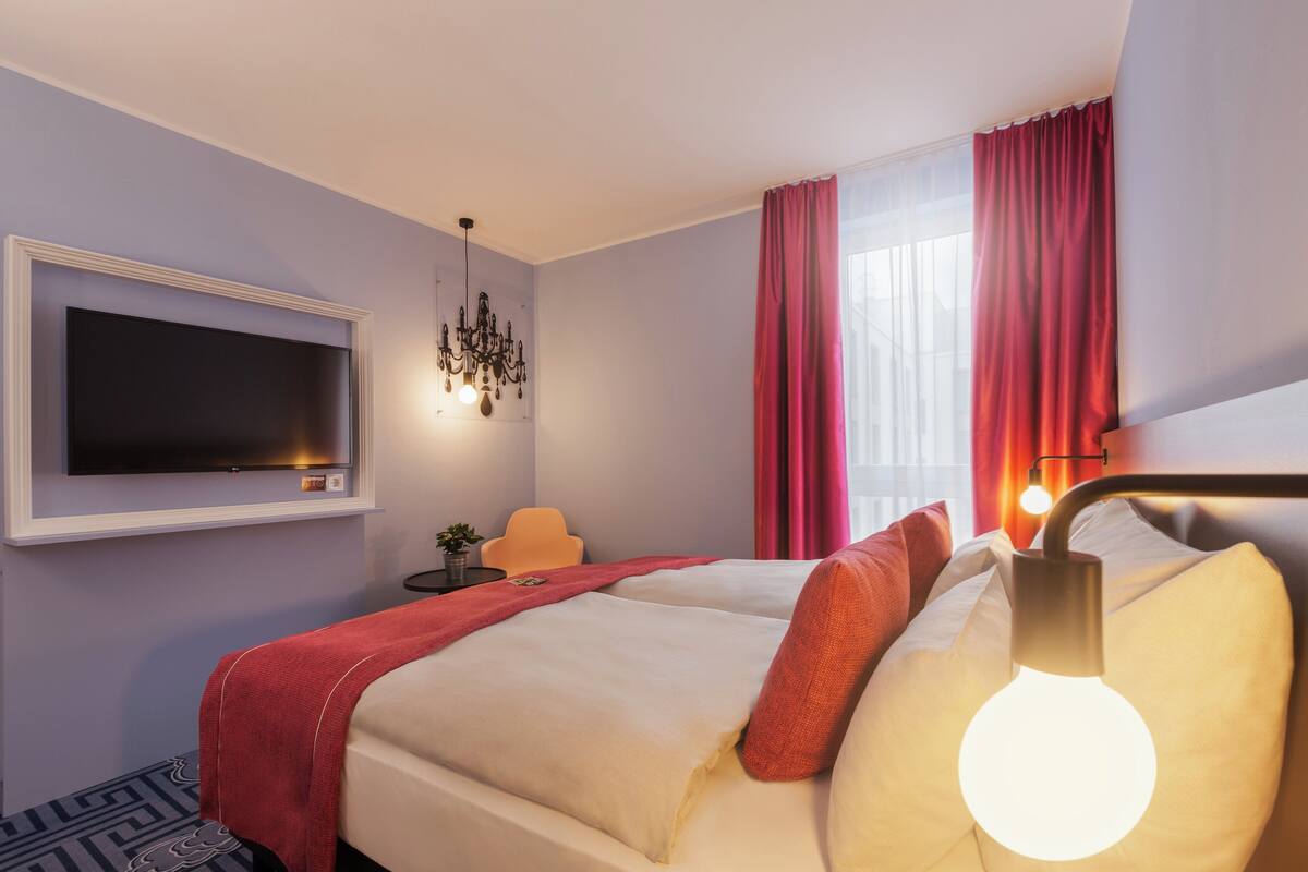 Ett hotellrum med en säng, en tv som är monterad på väggen, en kristallkrona och ett fönster med gardiner.
