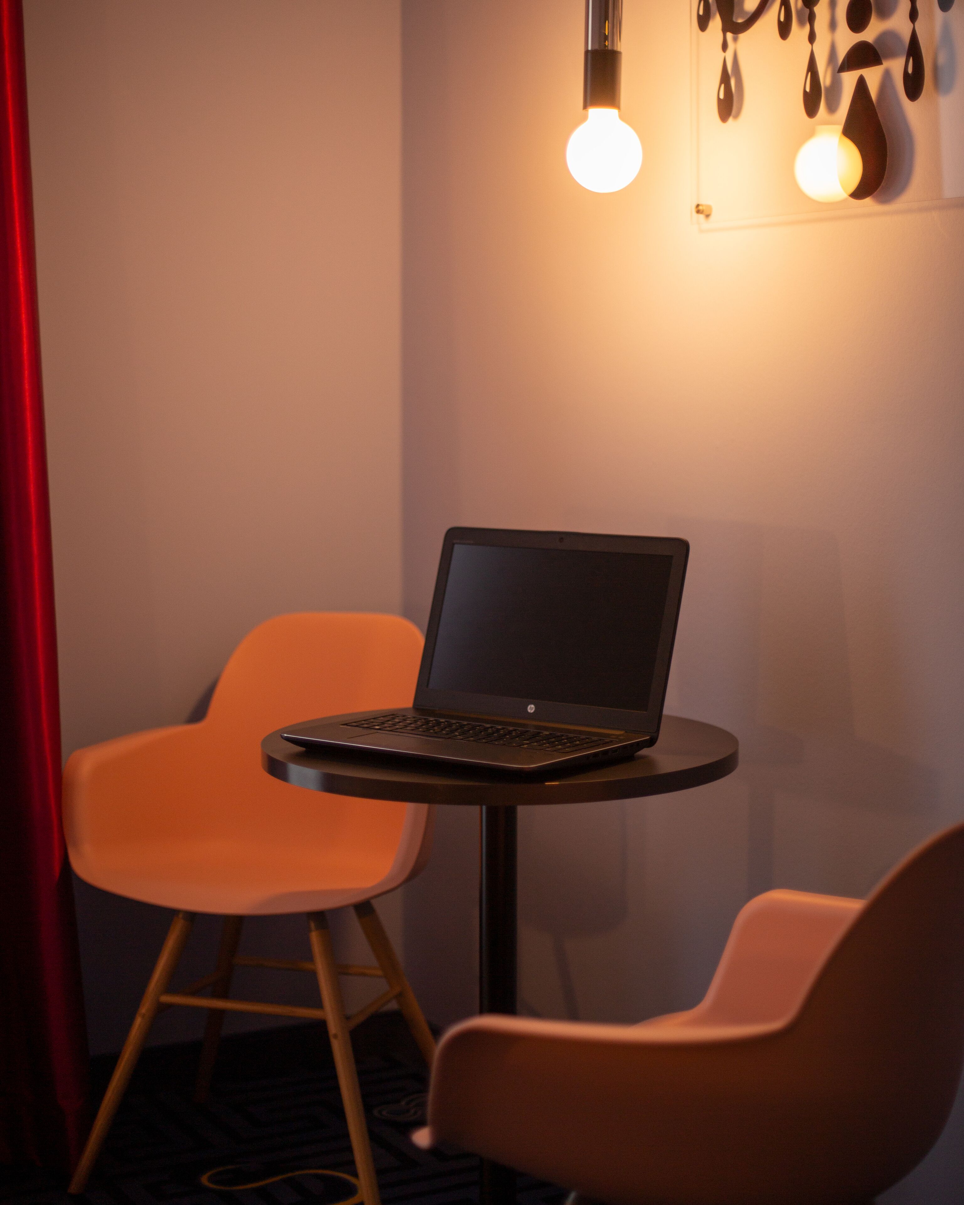 A laptop on a small round table with two orange chairs, a red curtain, and a wall with hanging light fixtures.