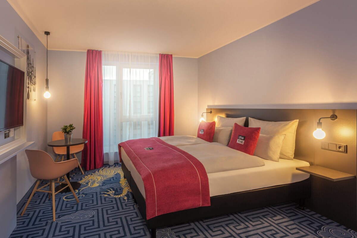 A hotel room with a bed, a desk, a chair, a TV, and a window with curtains.