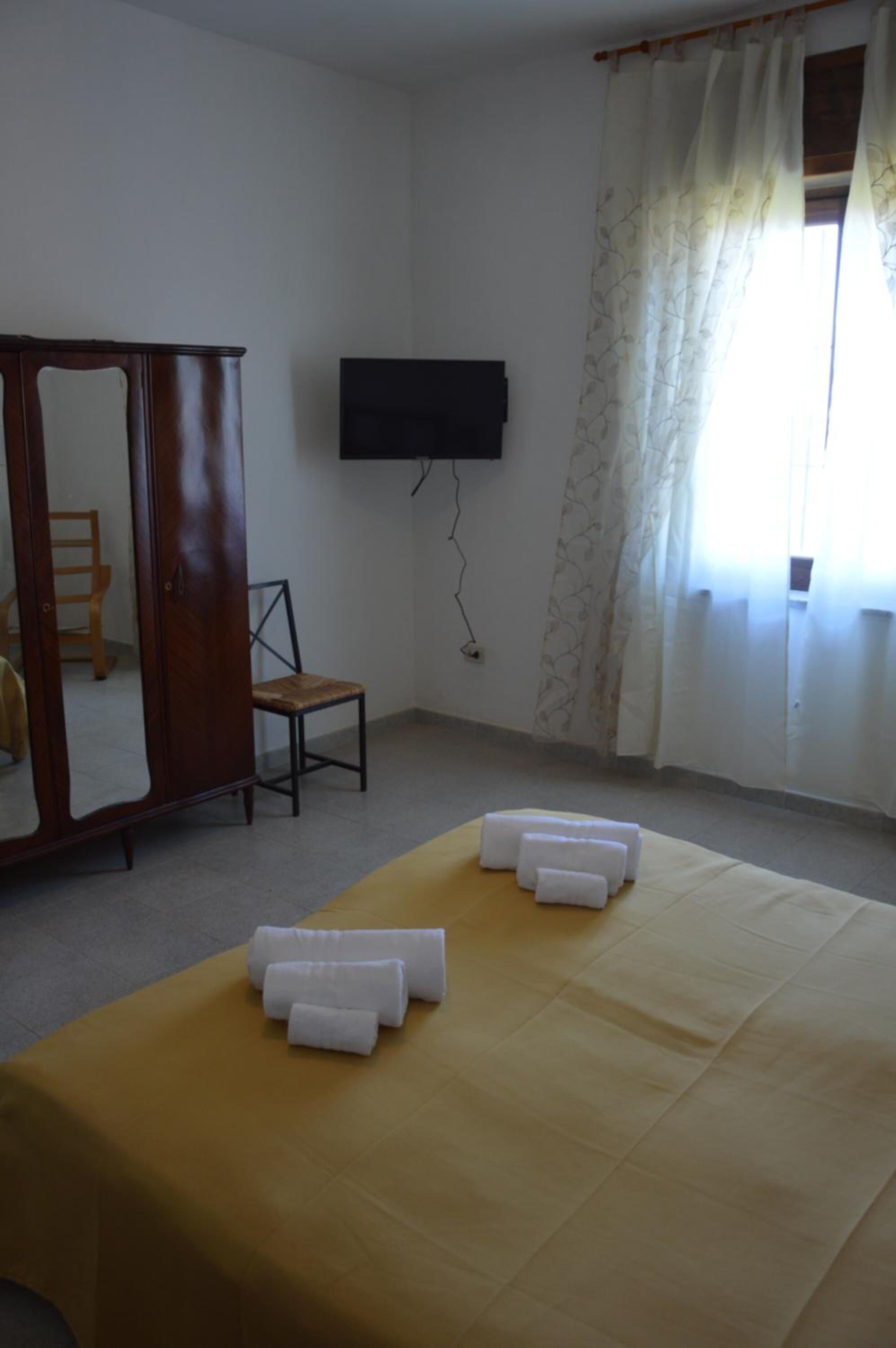A room with a bed, a wooden wardrobe, a chair, a television mounted on the wall, and a window with curtains.