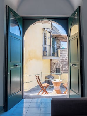 Puerta abierta que da a un balcón con mesa, sillas y vistas de un edificio de fachada de piedra.