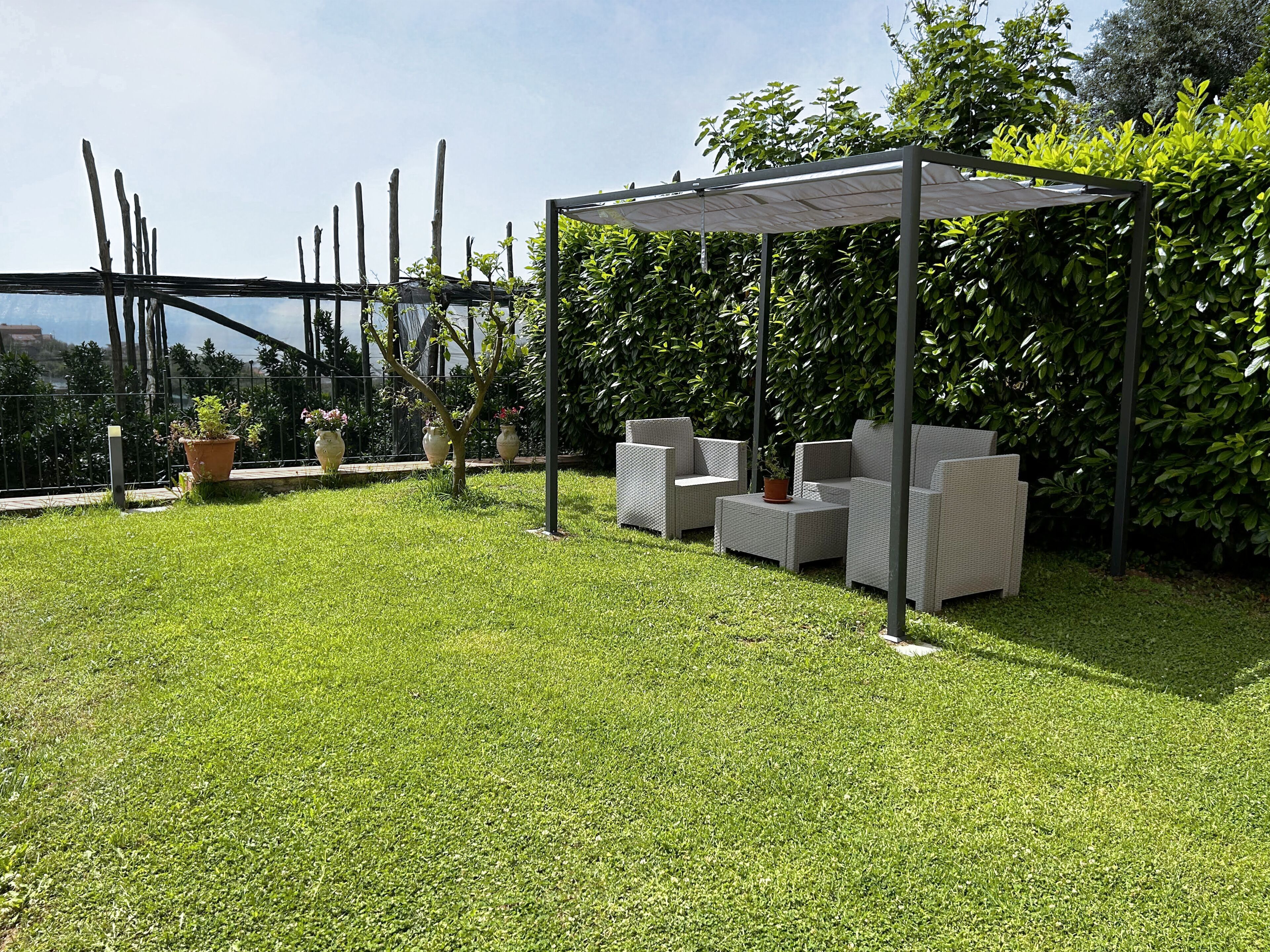 A patio area with outdoor furniture under a pergola, surrounded by lush greenery and potted plants.