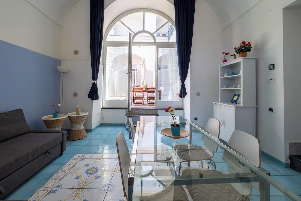 Uma sala de estar com uma mesa de vidro, cadeiras, um sofá, piso com azulejos azuis e brancos, uma grande janela em arco com cortinas e um armário branco com prateleiras.