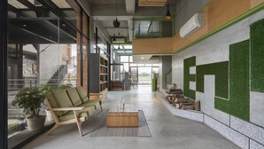 Een ruime lobby met een groene bank, een houten bankje en een bijzettafeltje. Er staan potplanten en er is een trap die naar een hoger niveau leidt.