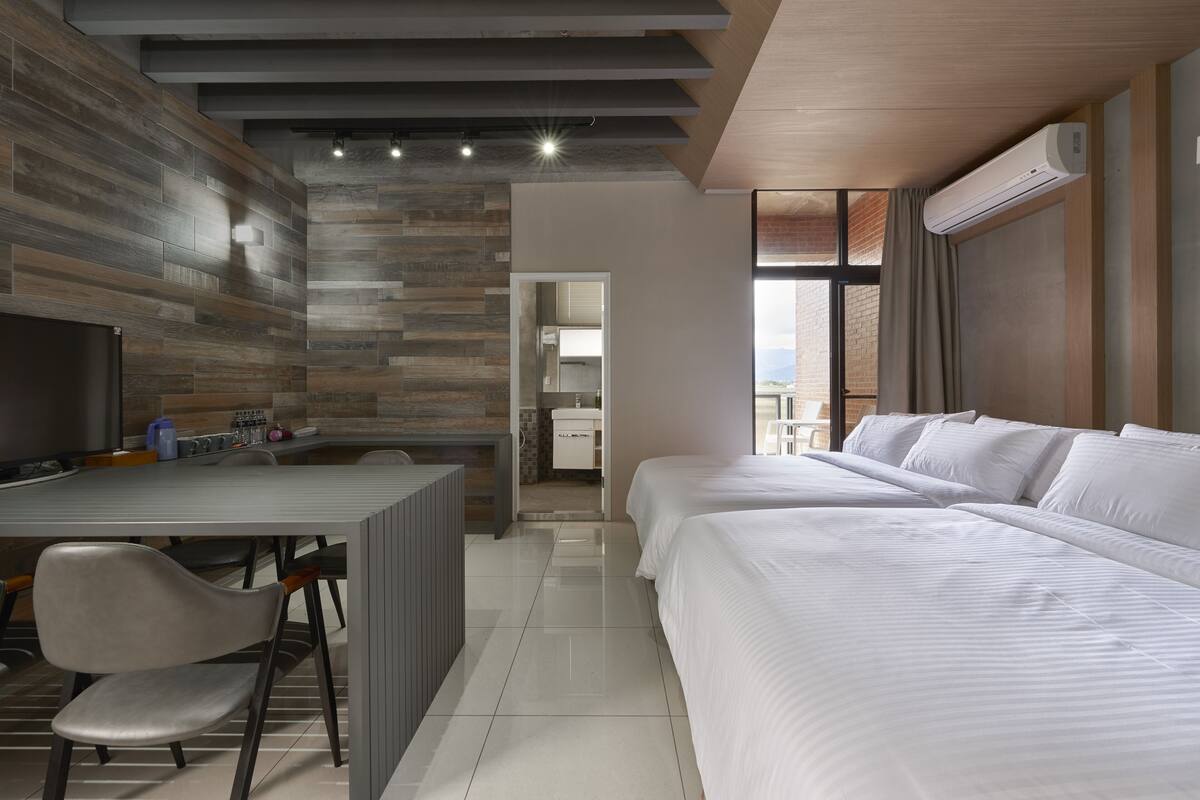 Una habitación de hotel moderna con una pared de madera, un escritorio con una silla, una cama grande y un baño visible a través de una puerta abierta.