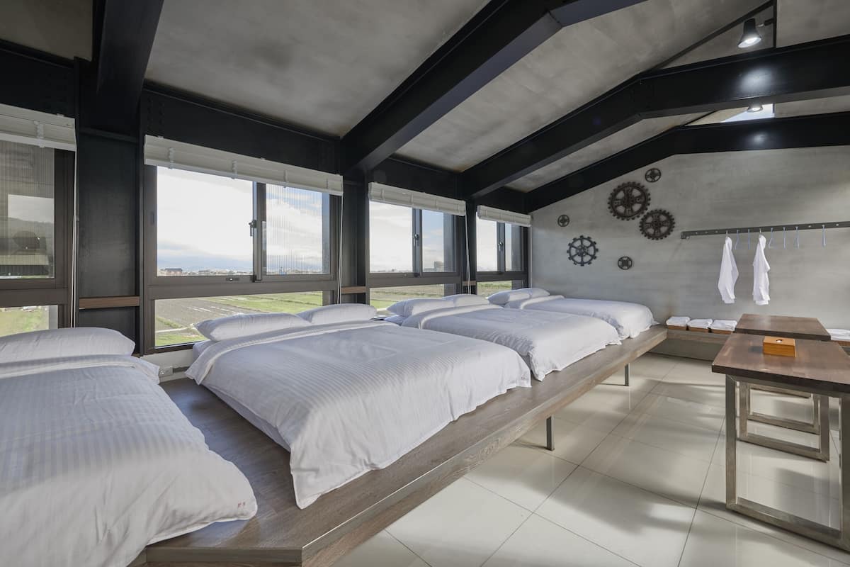 Una habitación de hotel moderna con ventanas amplias, una cama con ropa blanca, un banco de madera y una mesita.