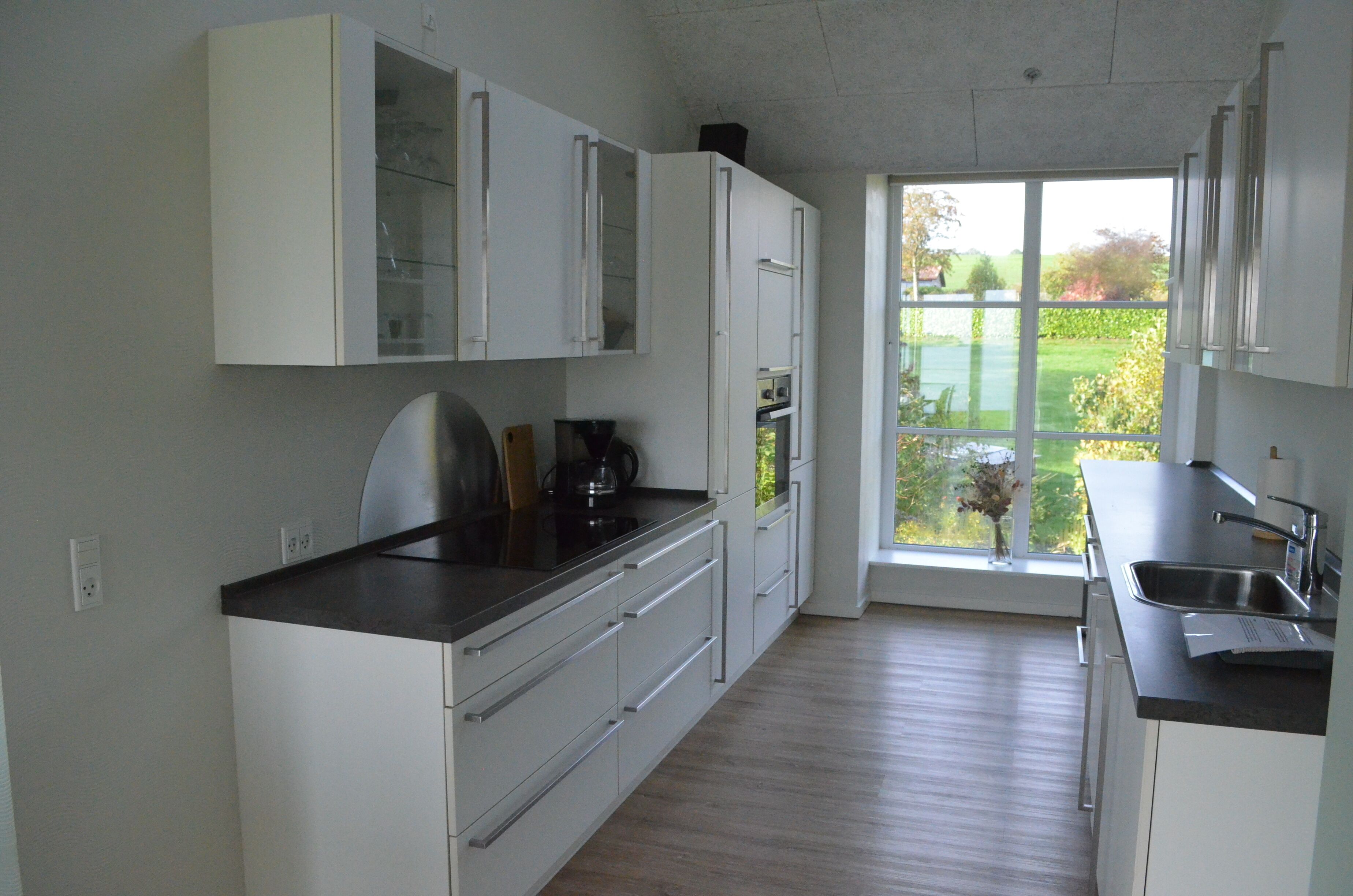 A modern kitchen with white cabinetry, a black countertop, and a view of a garden through a large window.