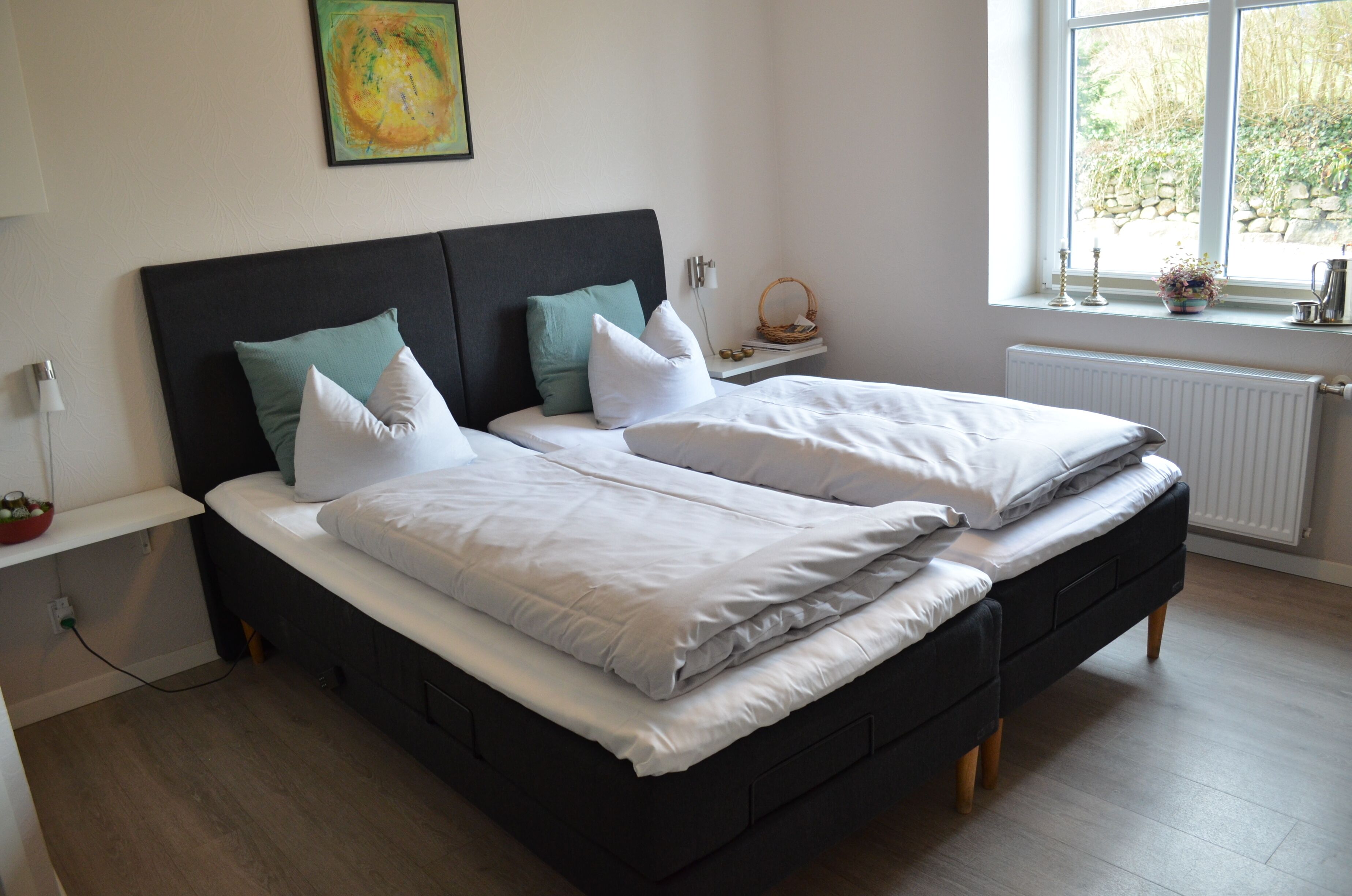 Cama doble con sábanas blancas, cabecera oscura y dos almohadas. Se observa una mesita de noche con una lámpara y un objeto decorativo pequeño. Una ventana muestra vista a zonas verdes.