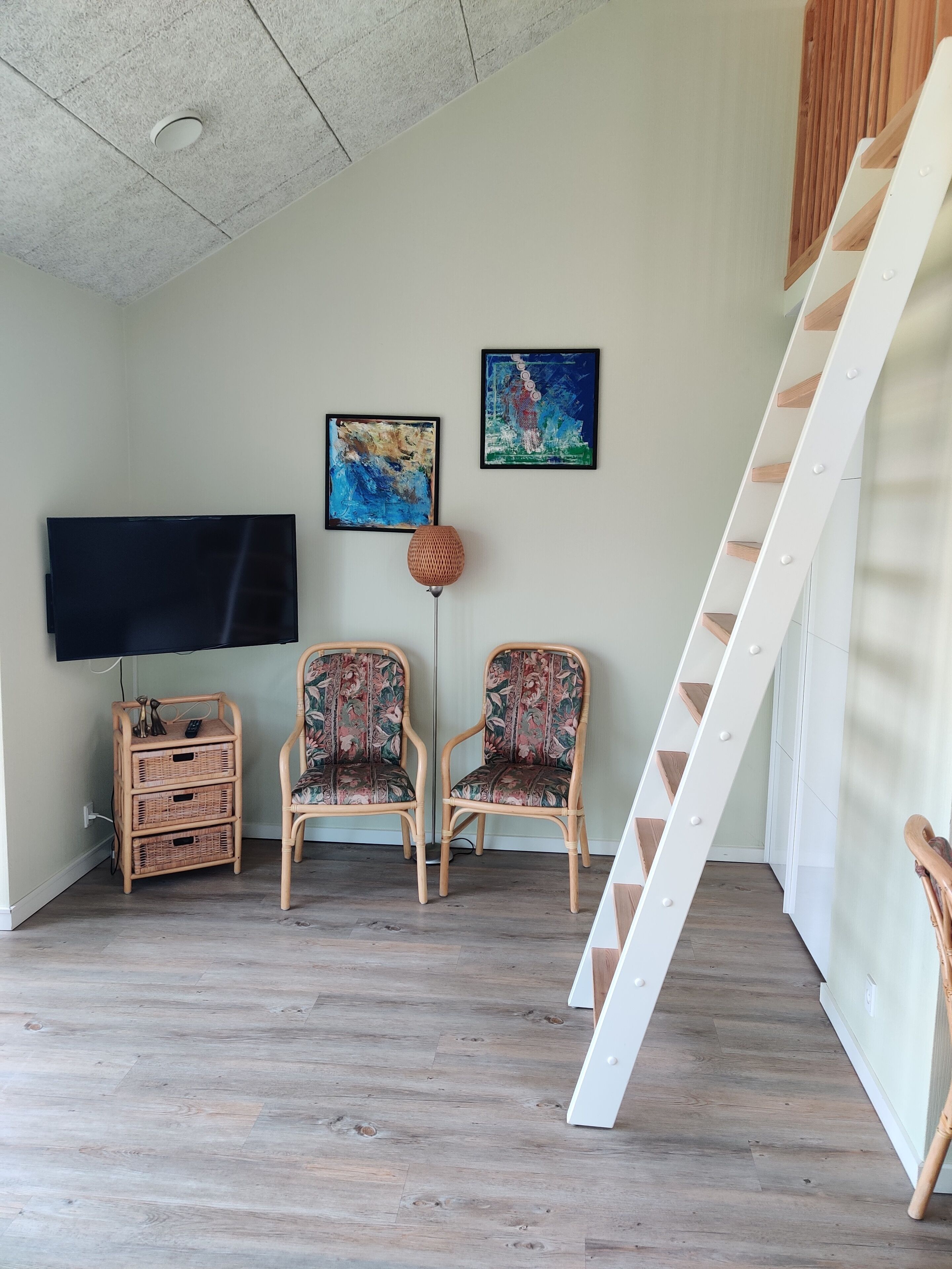 A room with a flat-screen TV, two wicker chairs, a wicker side table, and a white staircase.