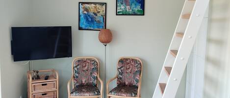 A room with a flat-screen TV, two wicker chairs, a wicker side table, and a white staircase.
