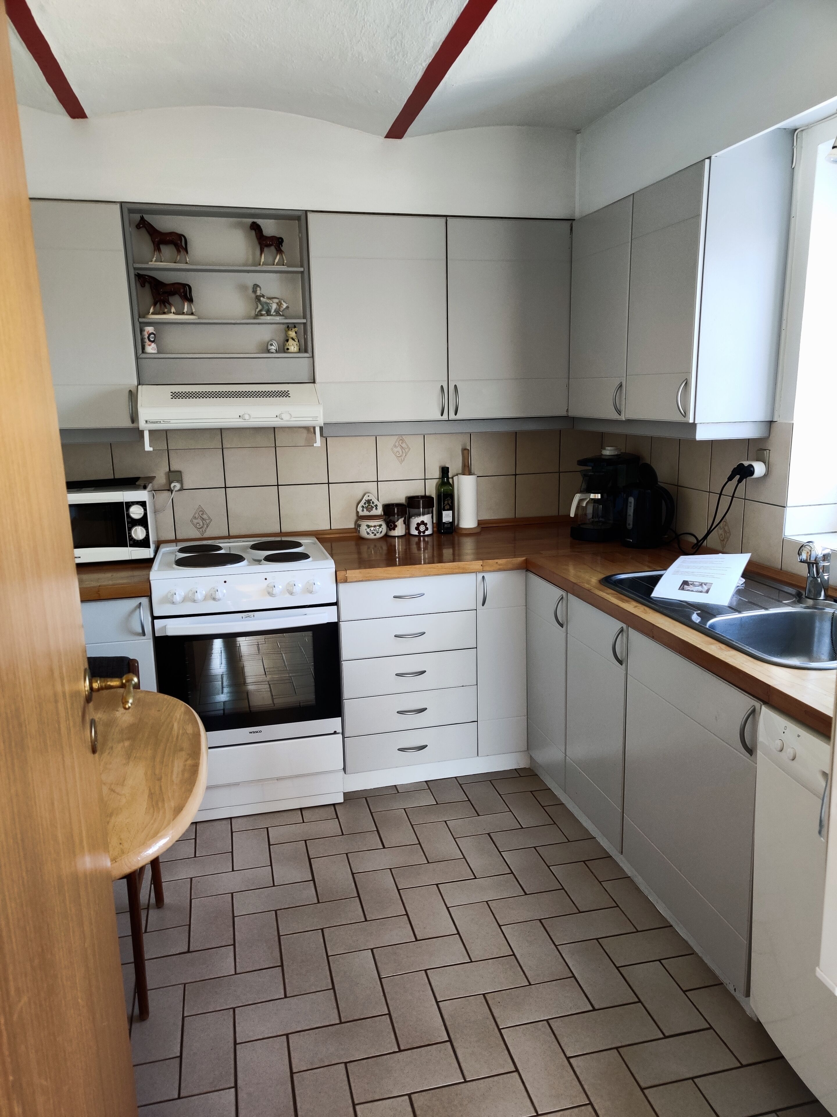 A kitchen with white cabinets, a stove, oven, microwave, and a sink. There are decorative items on the shelves and a wooden chair.