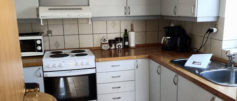 A kitchen with white cabinets, a stove, oven, microwave, and a sink. There are decorative items on the shelves and a wooden chair.