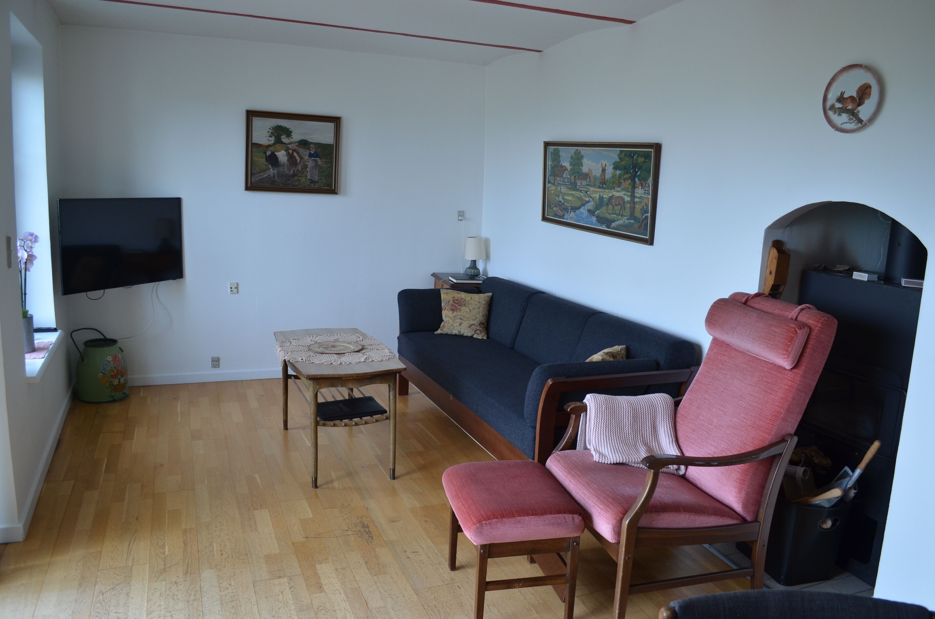 A living room with a sofa, a chair, a small table, and a television. There are two paintings on the wall and a window with a view of the outside.