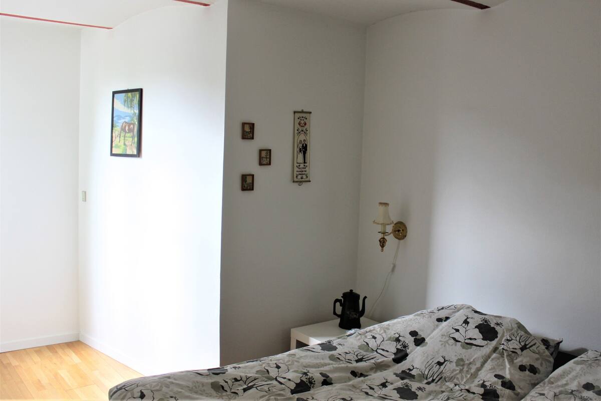 A bedroom with a bed, a bedside table with a kettle, a wall-mounted lamp, and a framed picture.