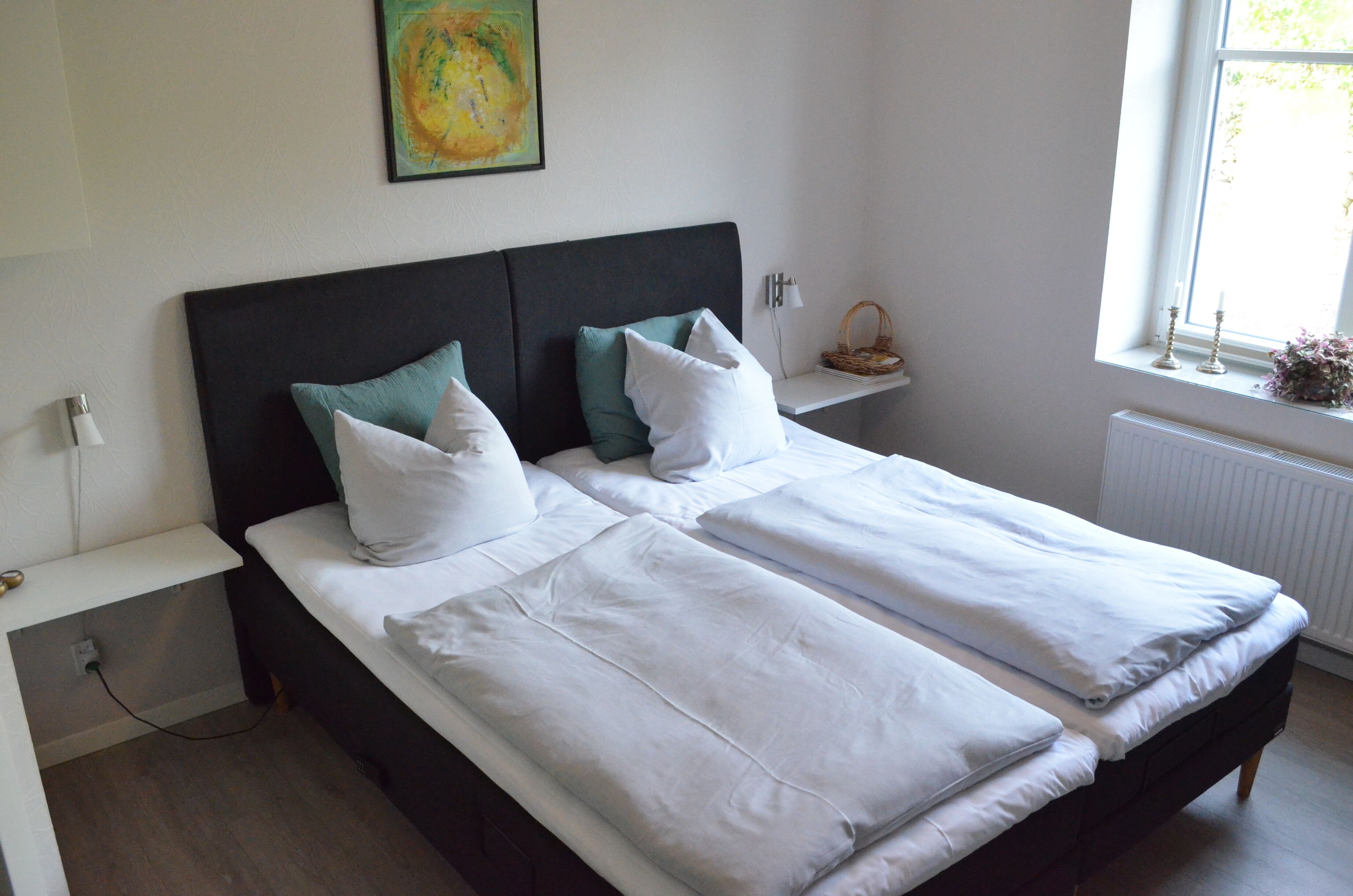 A double bed with white linens and green pillows, a bedside table, a small shelf with a basket, and a window with a view of greenery.