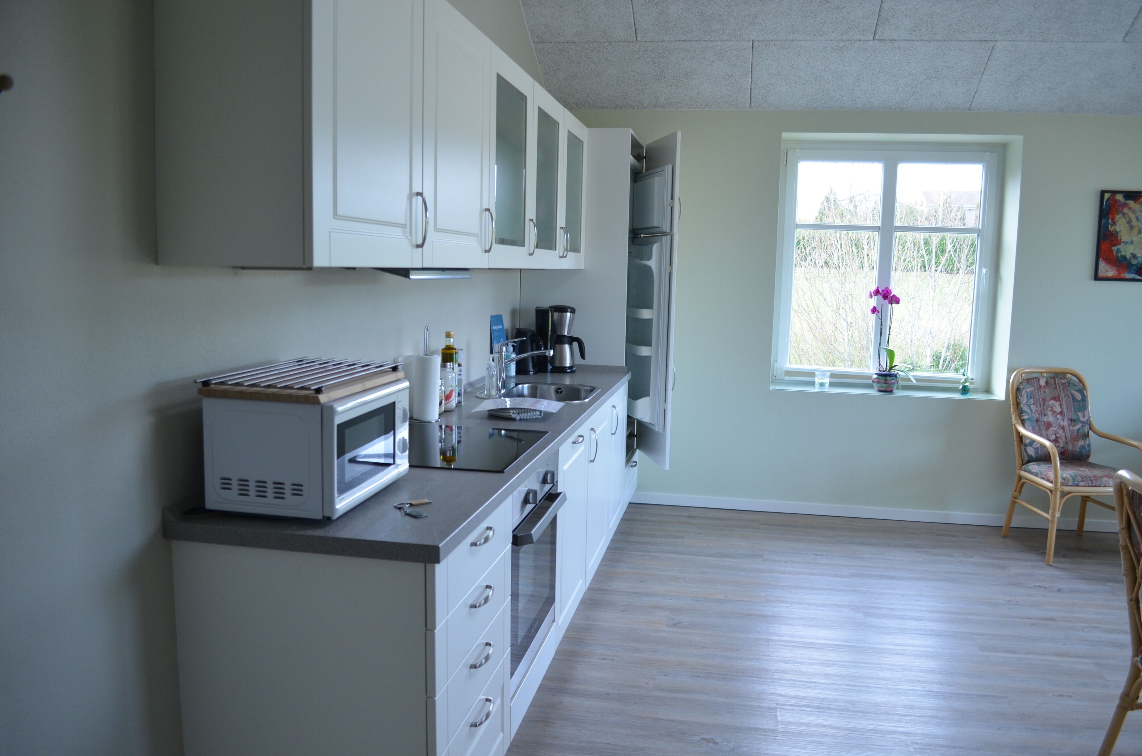 A kitchen with white cabinets, a microwave, and a coffee maker. There is a window with a view of trees and a chair near it.