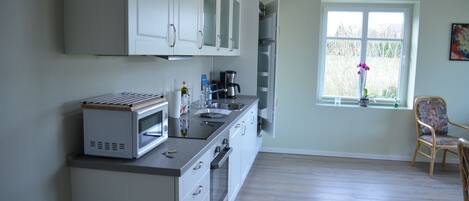 A kitchen with white cabinets, a microwave, and a coffee maker. There is a window with a view of trees and a chair near it.