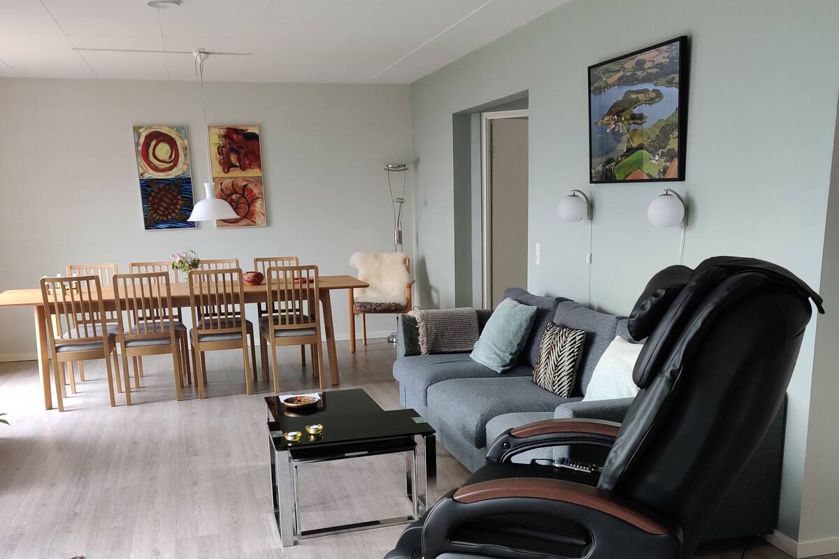A modern living room with a grey sofa, a massage chair, a dining table with chairs, and a painting on the wall.