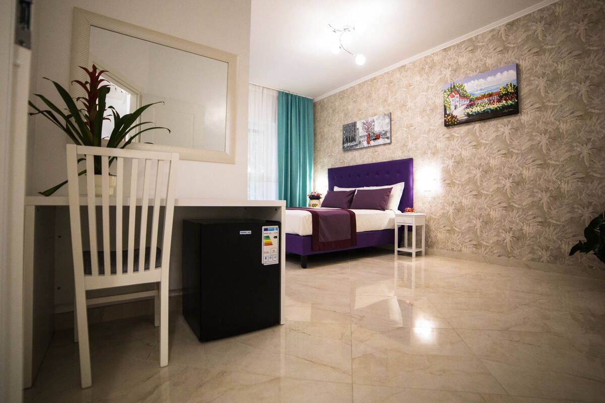 A hotel room with a bed, a small desk, a chair, a refrigerator, and a painting on the wall.