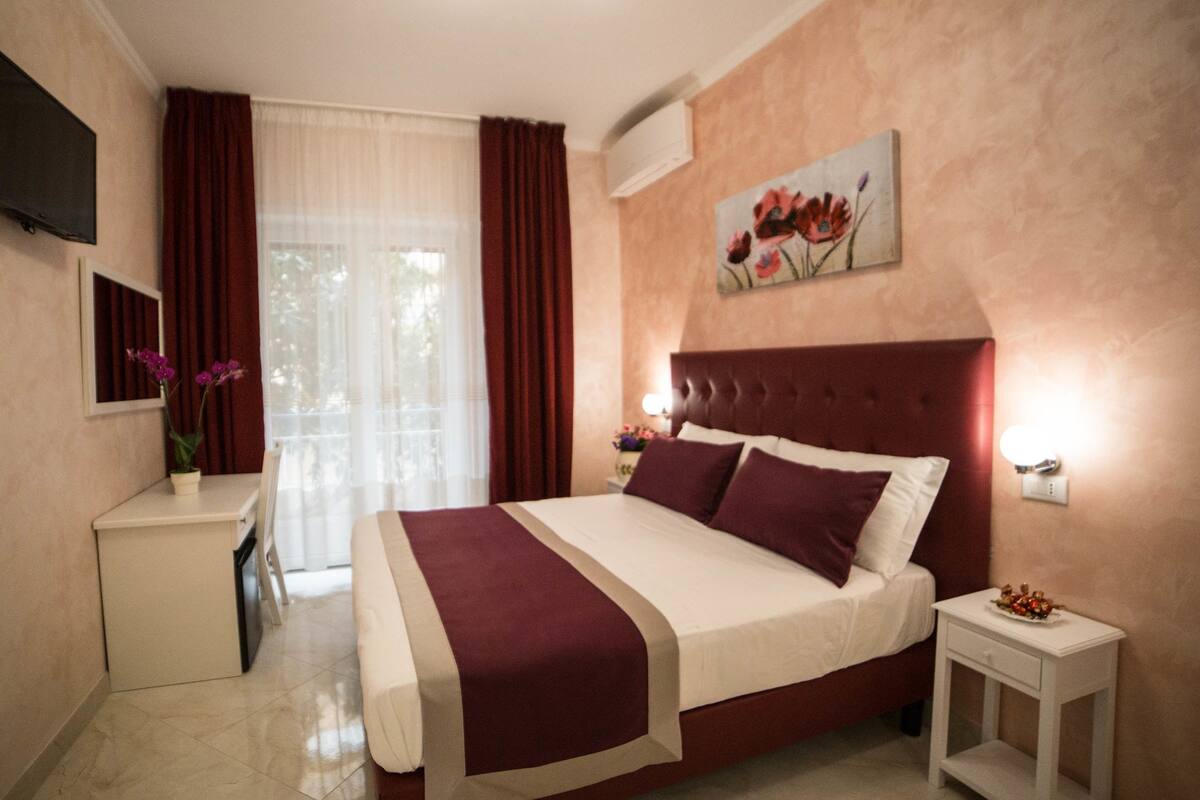 A hotel room with a large bed, a bedside table, a TV mounted on the wall, and a window with curtains.