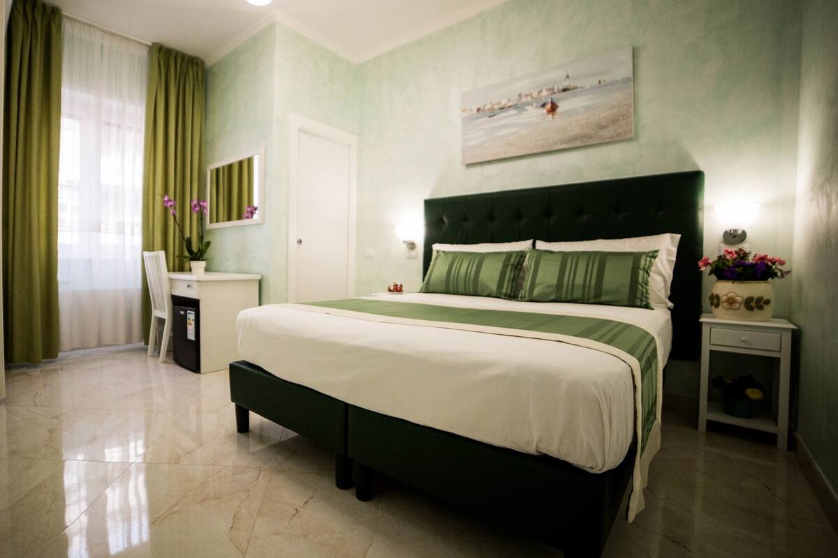 A bedroom with a bed, a desk, a chair, a nightstand, and a painting on the wall.
