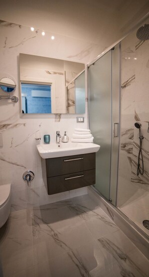 A modern bathroom with a marble floor, a glass shower enclosure, a white sink with a dark cabinet, and a mirror above the sink. - St. Peter Area Luxury Suites (Rome)