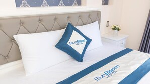Habitación de hotel con cabecera capitonada, cama con ropa de cama blanca y azul, almohada con el logotipo del Hotel SunBeach y una mesita de noche con un florero.