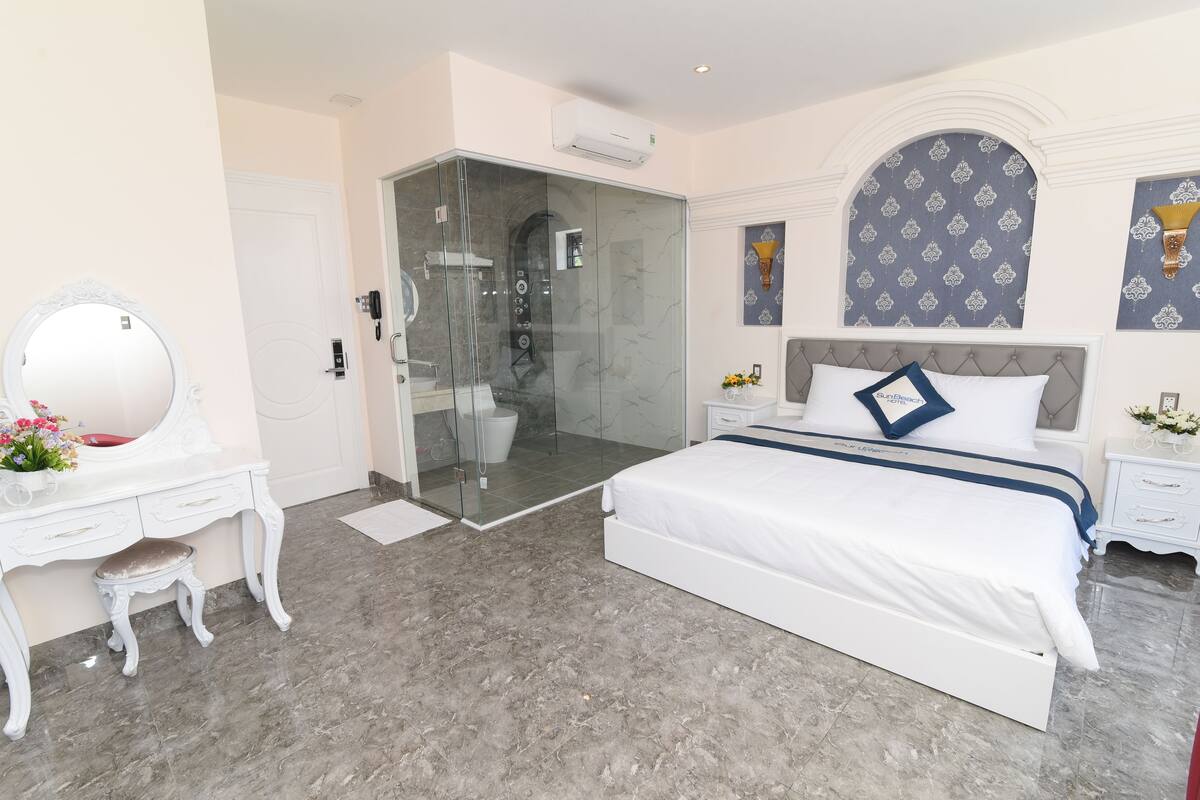 A modern bedroom with a glass-enclosed shower, a white bed with a blue accent pillow, and a white vanity with a mirror.