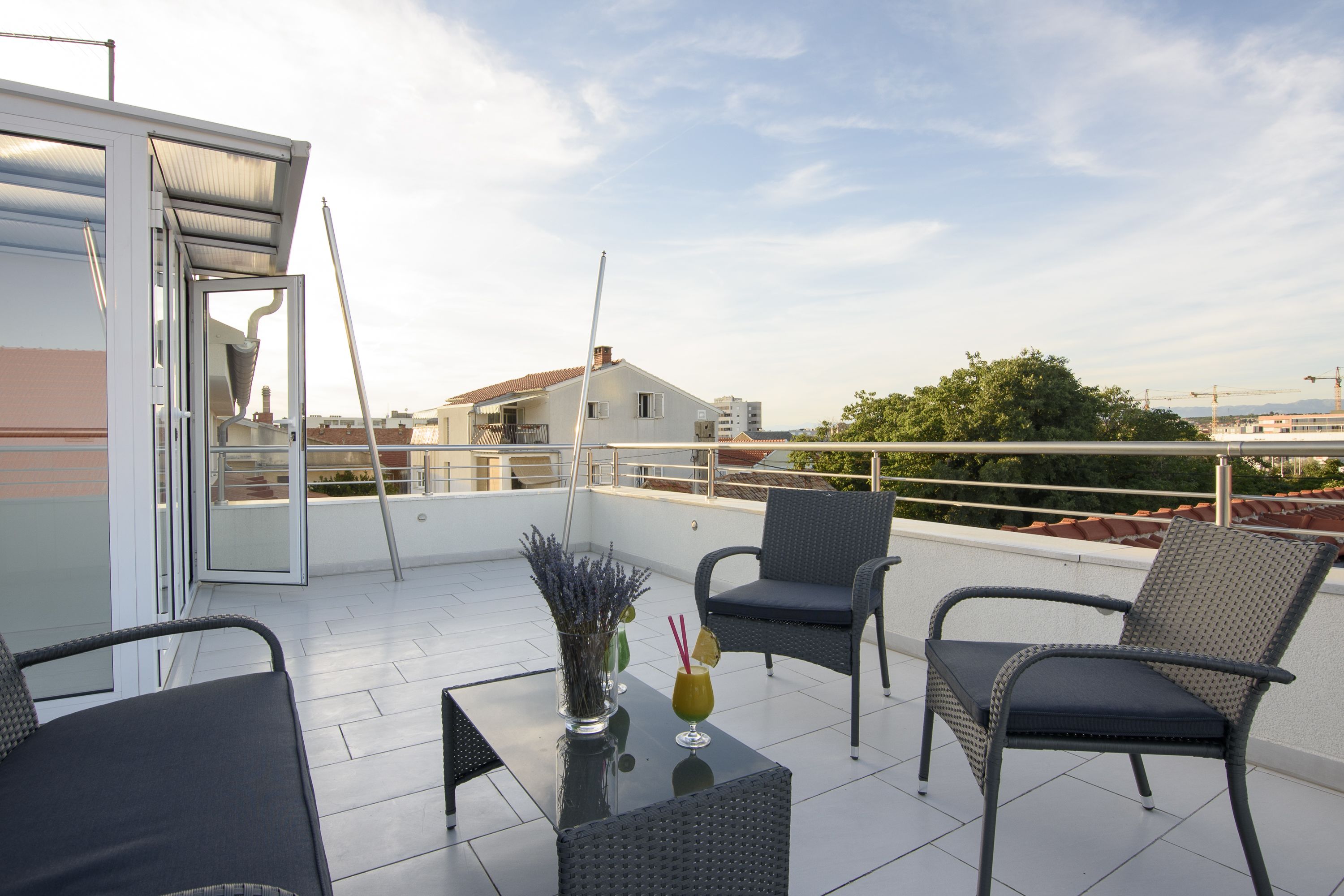 A rooftop terrace with a glass door, two wicker chairs, a small table with a vase and drinks, and a view of buildings and trees.
