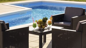 A poolside setting with wicker furniture, a fruit bowl, and two drinks.