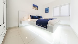 A modern bedroom with a white bed, blue bedding, and a large window with blinds. - Holiday Home Apartments Bellamare (Zadar)