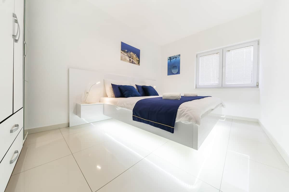 Quarto moderno com cama branca, roupa de cama azul e uma janela grande com persianas.