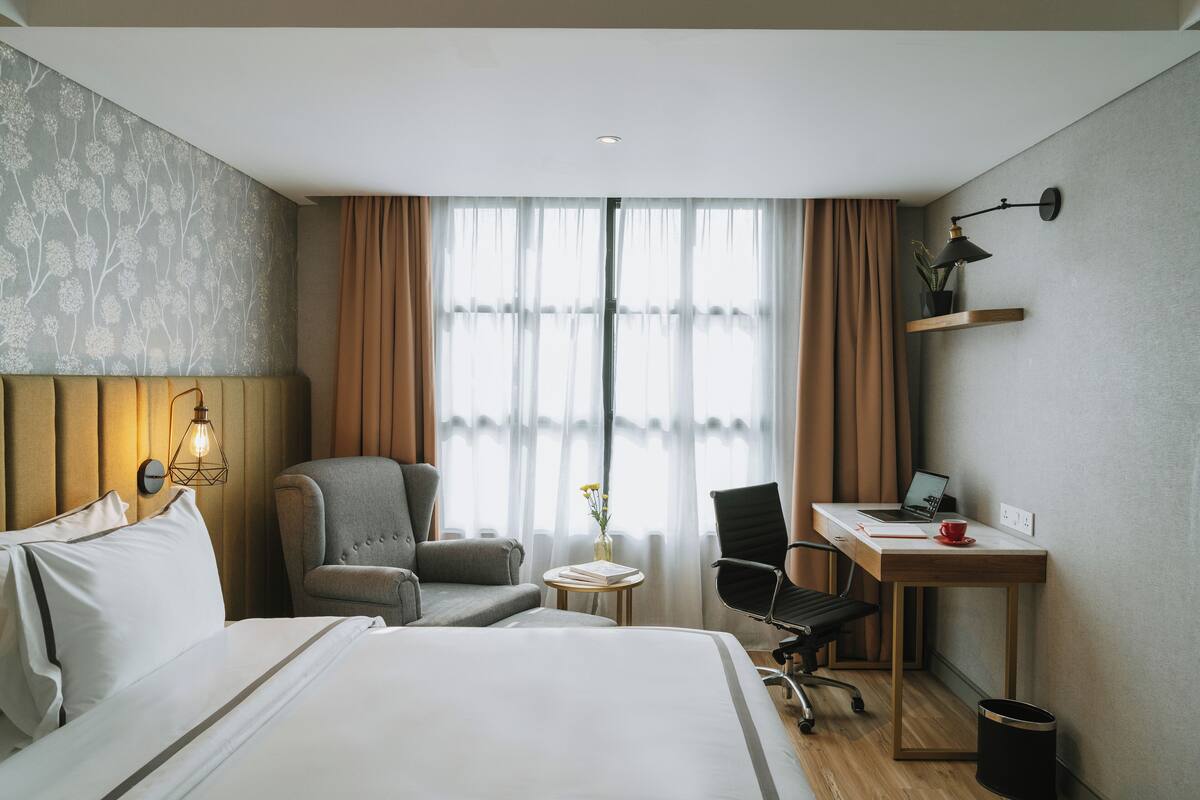 A hotel room with a bed, two chairs, a desk with a laptop, a small table with a vase, and a wall-mounted lamp.