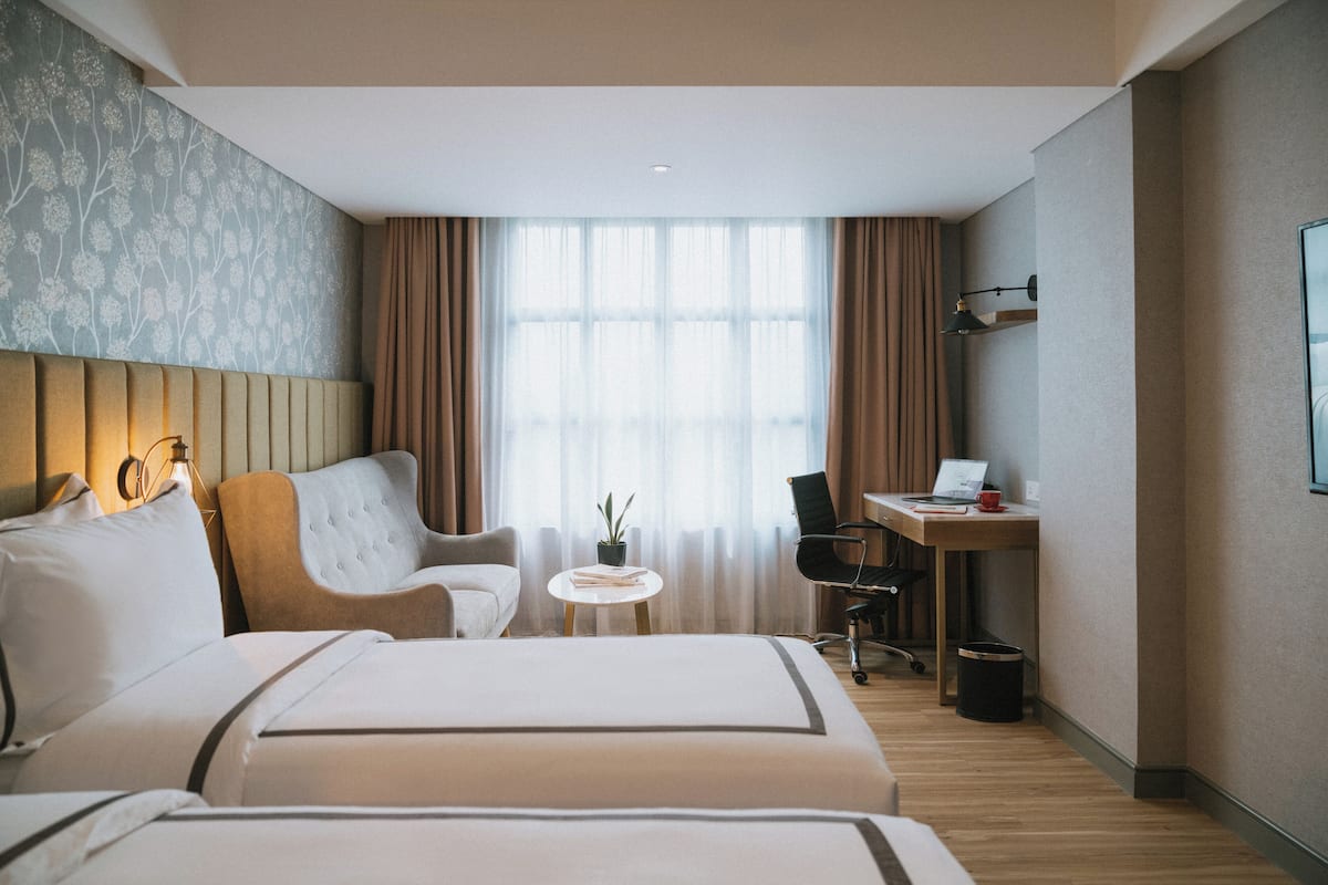 A hotel room with two beds, a desk, a chair, a small table with a plant, and a window with curtains.