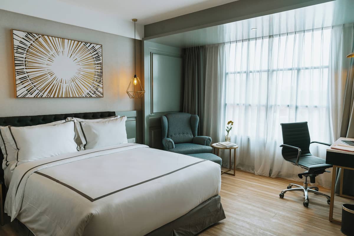 A hotel room with a large bed, a desk with a chair, a small side table, and a large window with curtains.
