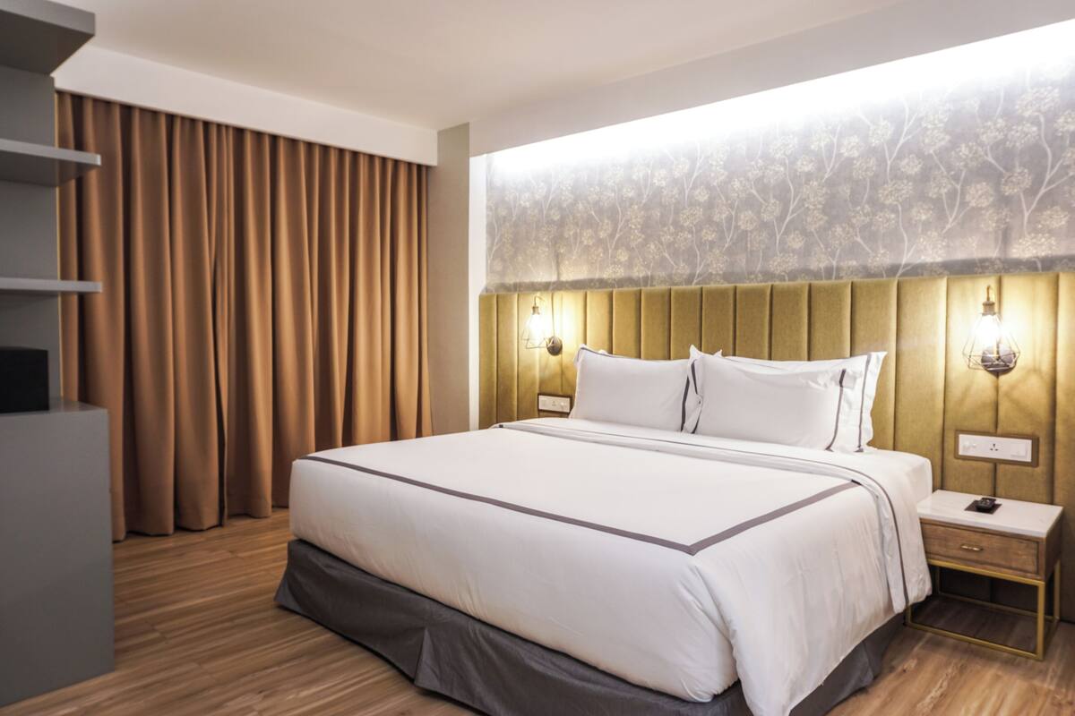 A modern hotel room with a large bed, wooden flooring, a headboard with wall-mounted lights, and a view of the outdoors through sheer curtains.