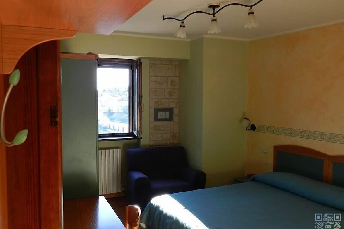A small, cozy room with a bed, a chair, and a table. There is a window with a view of a building outside.