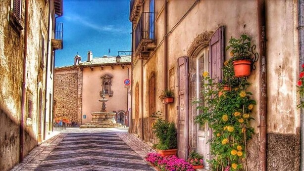 A narrow cobblestone street lined with buildings, a fountain, and flower boxes. - Garnì B&B La Rua-Pescocostanzo (Pescocostanzo)