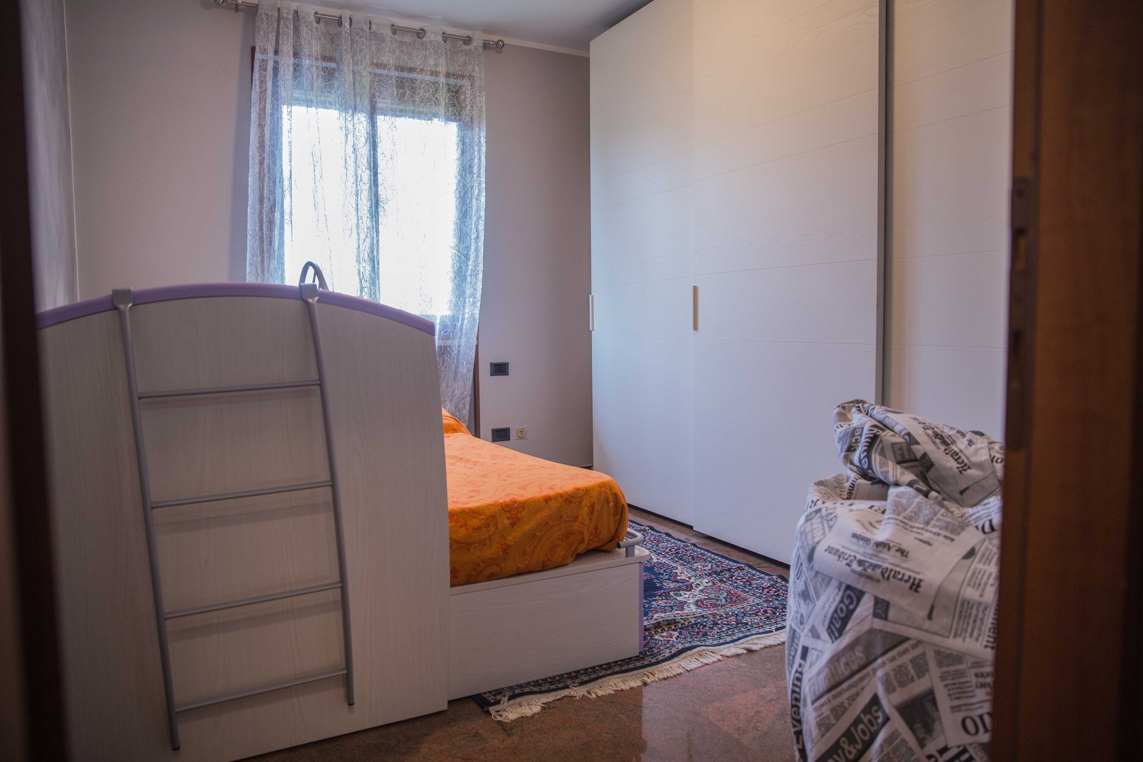 A compact bedroom with a bunk bed, a window with sheer curtains, a patterned rug, and a door with a newspaper print.