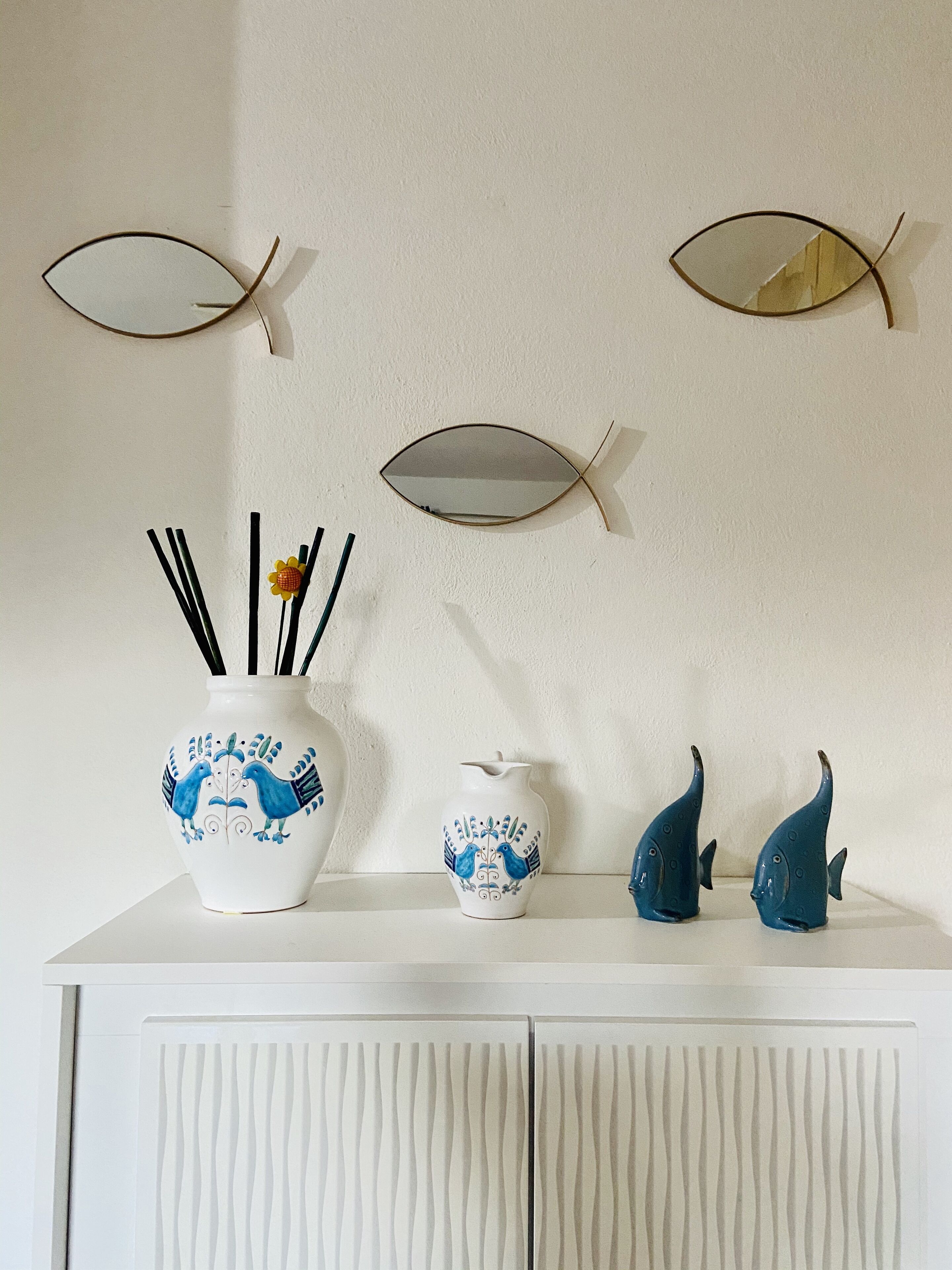 Ein weißes Schranksystem mit einer weißen Vase, einem kleinen weißen Krug und zwei blauen, fischförmigen Vasen darauf.