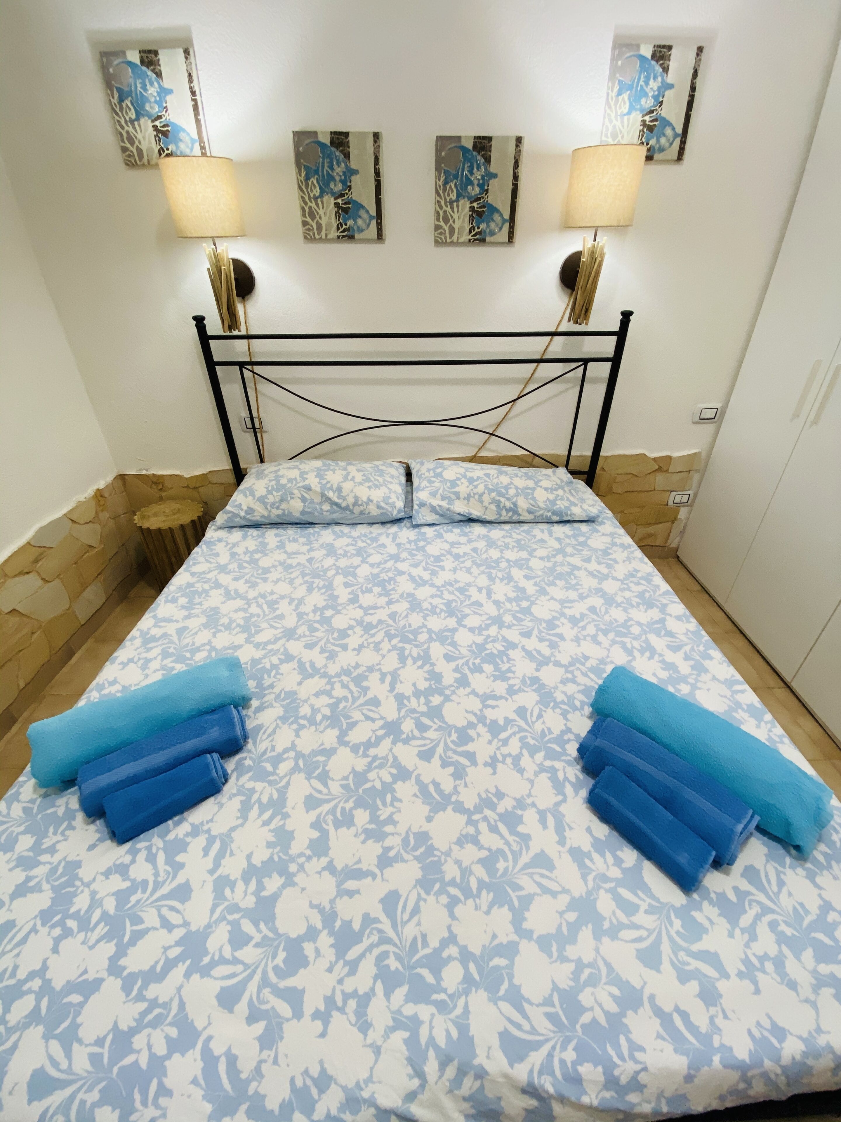 A bed with a floral-patterned bedspread, two rolled towels, and two wall-mounted lamps.