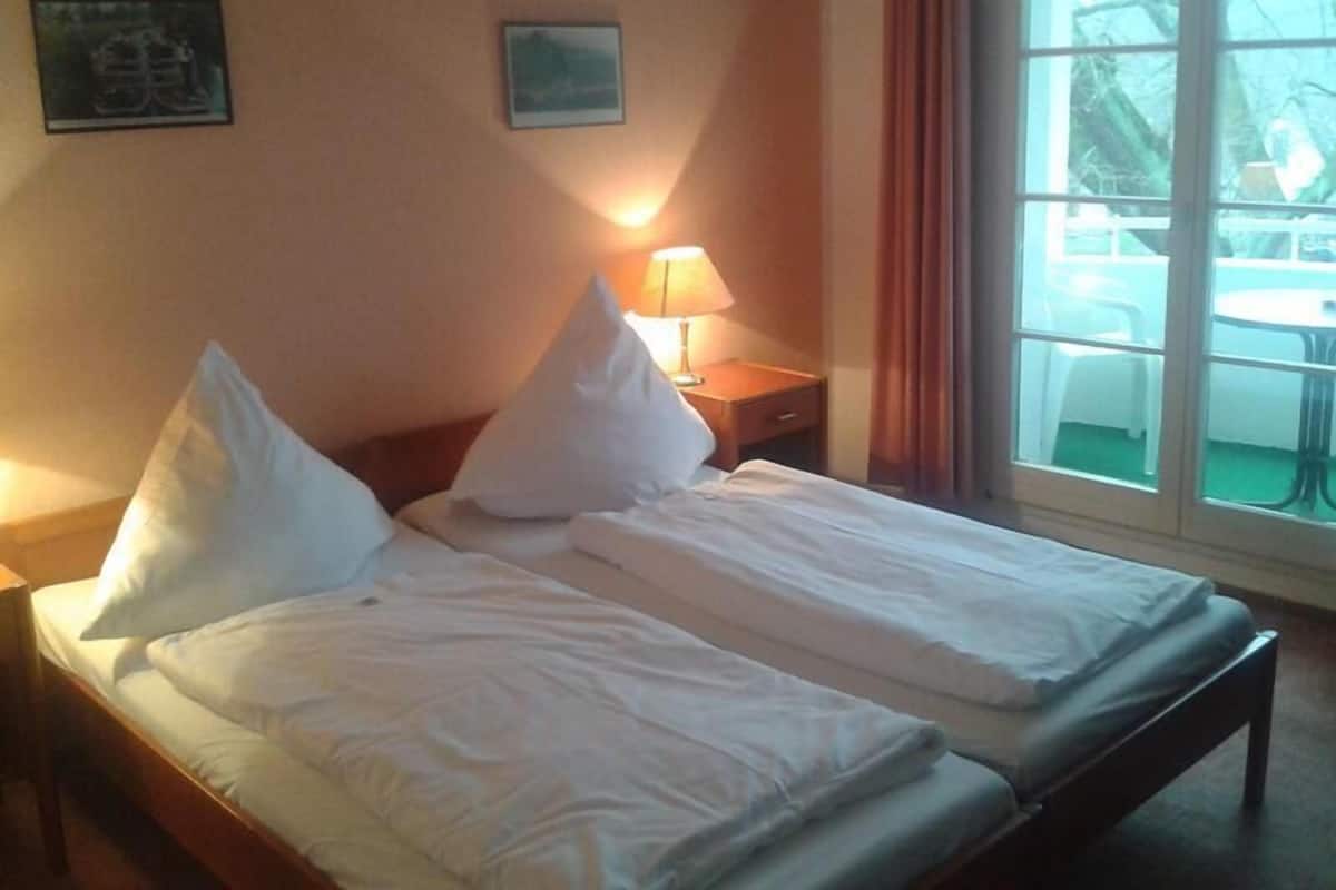 Ein Hotelzimmer mit zwei Betten, einem Nachttisch mit Lampe und einem Fenster mit Blick auf Gartenmöbel.