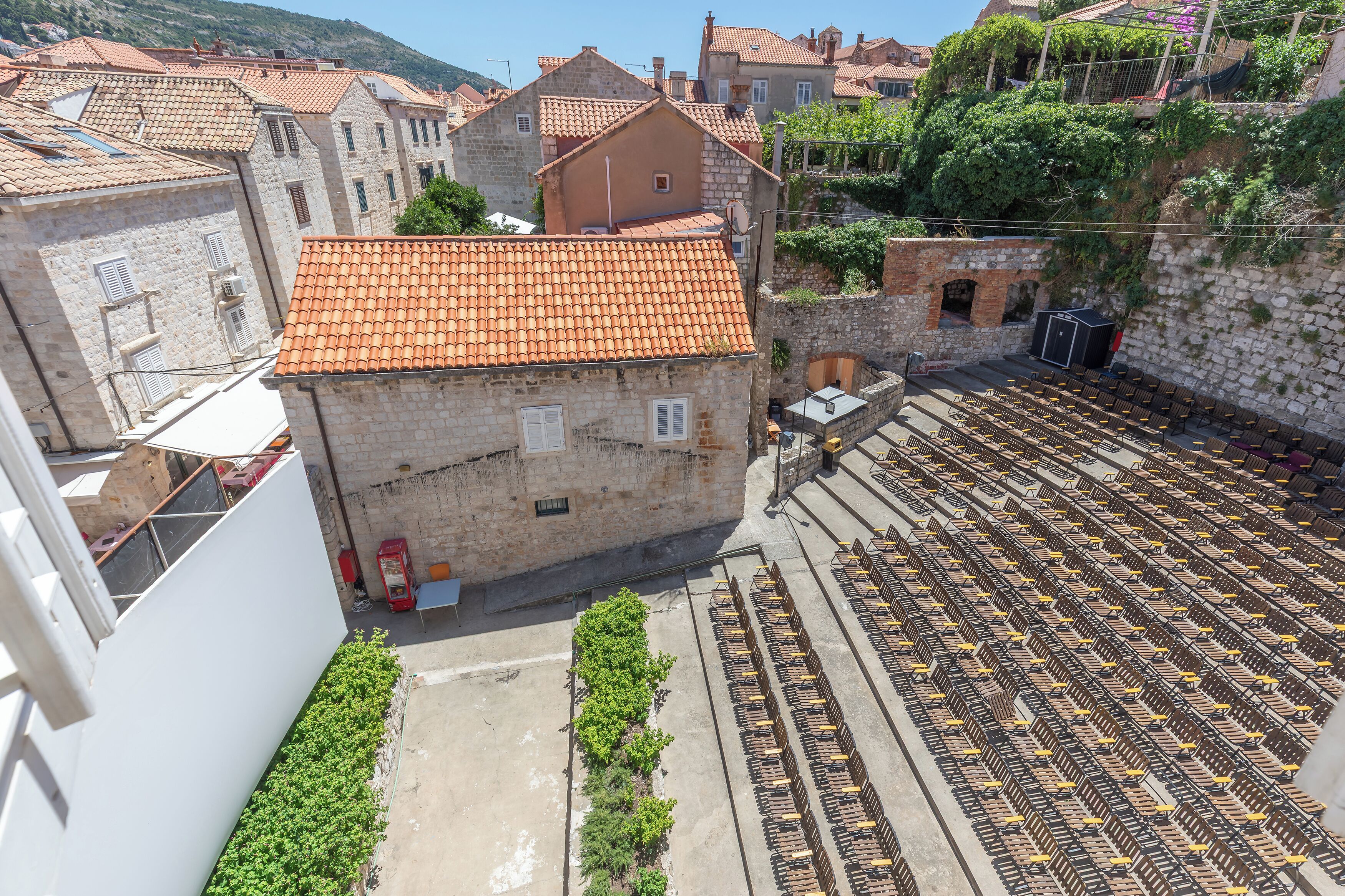 A stone building with a tiled roof, a courtyard with rows of parked cars, and a view of surrounding buildings.